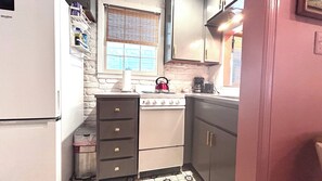 A compact kitchen with a white refrigerator, a stove, and a sink. There is a window with blinds, and a shelf with various items. - Welcome to the Nest, a unique private carriage house--Great Value! (Savannah)