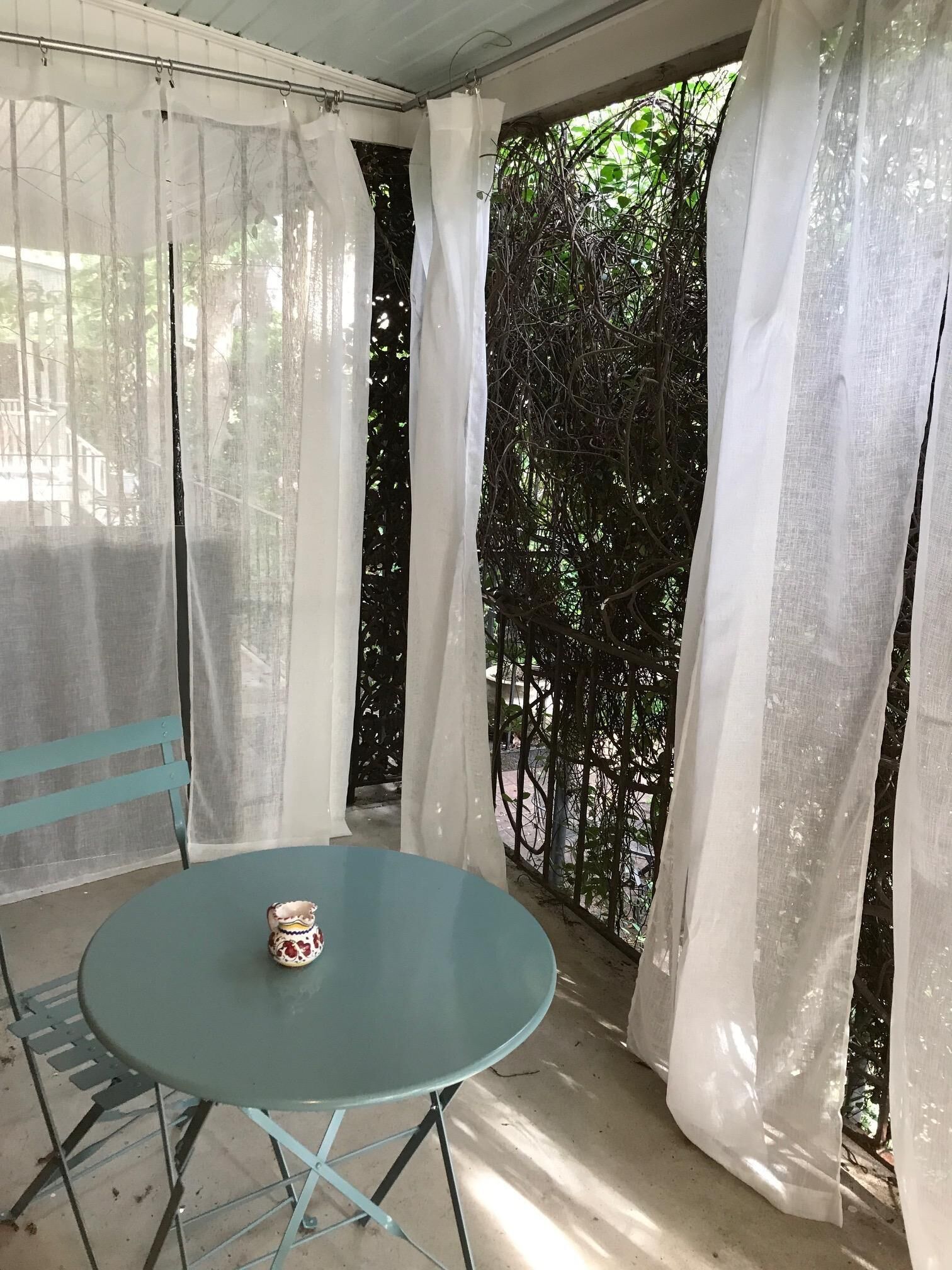 A small, round table with a single decorative item on it, situated in a covered outdoor area with white curtains and a view of greenery.
