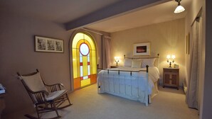 A bedroom with a bed, a rocking chair, a nightstand with lamps, and a stained glass window. - Downtown Port Townsend - Hillside Penthouse Vacation Rental (Port Townsend)