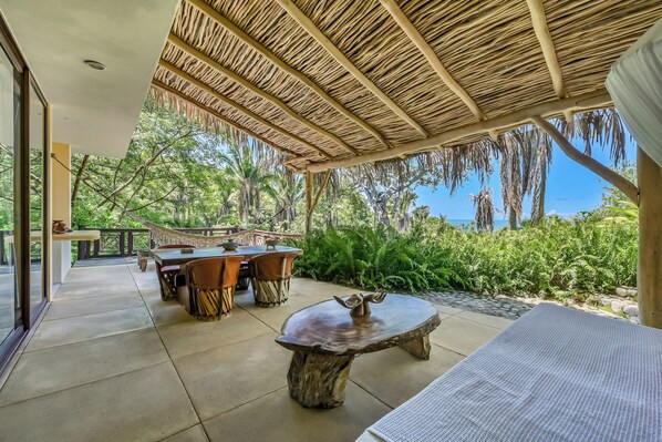 An outdoor patio with a thatched roof, a round wooden table, and a dining table with chairs.