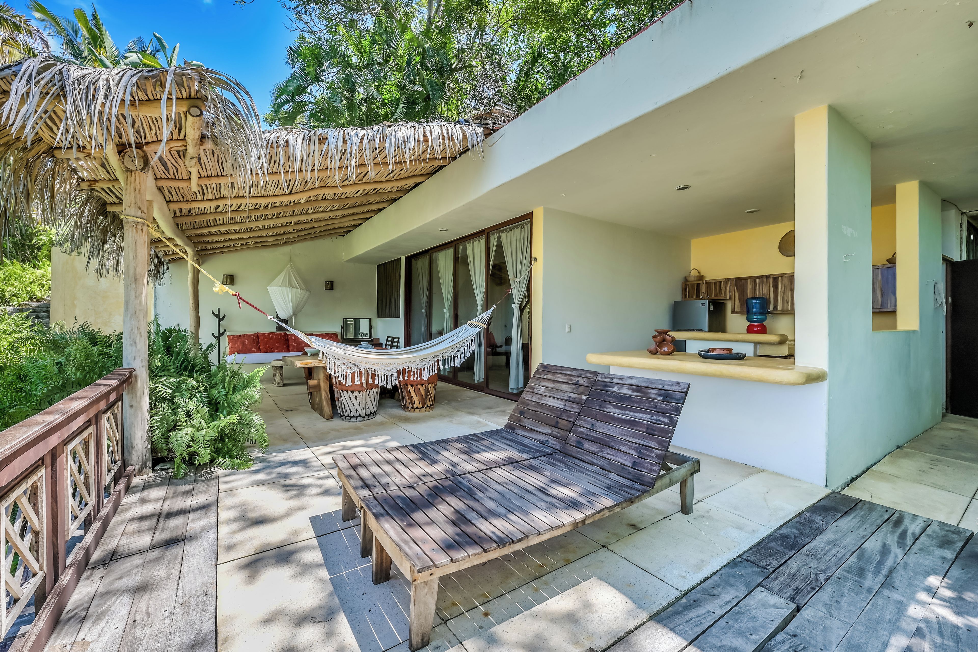 Un banco de madera y una hamaca en un patio con techo de paja, una zona de cocina y vistas al follaje circundante.