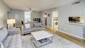 Un salon moderne avec un canapé gris, une table basse blanche et un tapis à motifs. On aperçoit une fenêtre avec des stores, un ventilateur de plafond et un lave-linge à l’arrière-plan.