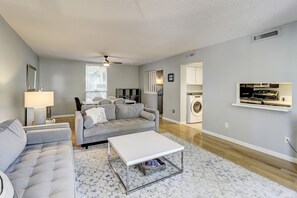 Ein modernes Wohnzimmer mit einem grauen Sofa, einem weißen Couchtisch und einem gemusterten Teppich. Ein Fenster mit Jalousien, ein Deckenventilator und eine Waschmaschine sind im Hintergrund zu sehen.