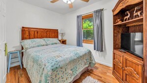 A bedroom with a wooden headboard, a bed with floral bedding, a ceiling fan, a window with curtains, and a wooden cabinet with a television.