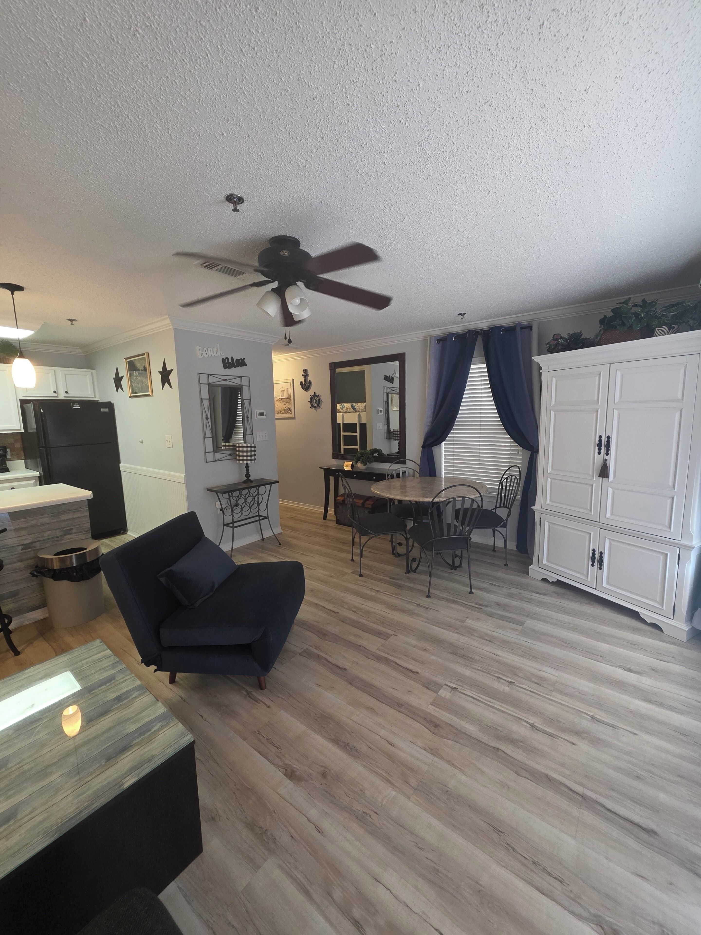 A modern living room with a dark blue sofa, a dining area with a round table and chairs, a kitchen area with a refrigerator, and a ceiling fan.