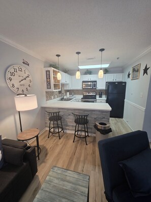 A compact kitchen with a central island, bar stools, a clock, pendant lights, and a refrigerator.