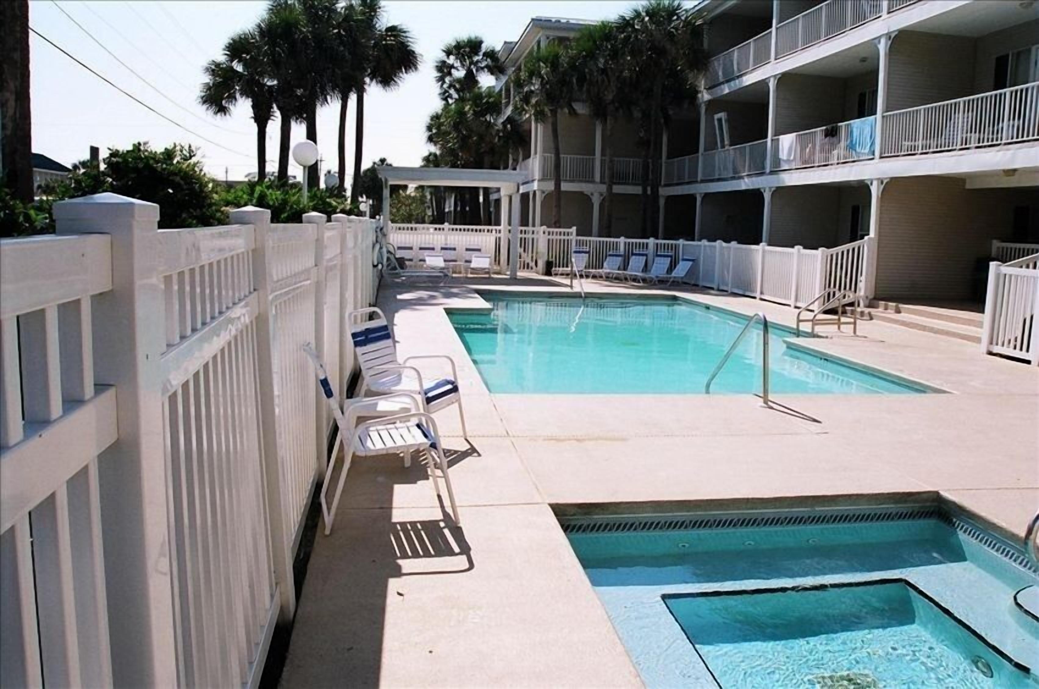 A swimming pool area with lounge chairs and a hot tub, surrounded by a white fence and palm trees.
