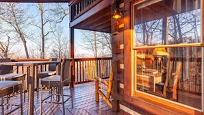 Una terraza de madera con mobiliario exterior y un amplio ventanal con vistas a un área boscosa.