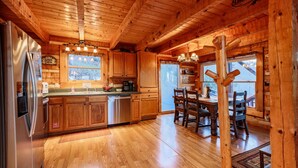 Interior de una cabaña de madera con cocina, que cuenta con alacenas de madera, un refrigerador de acero inoxidable y un comedor con mesa y sillas de madera.
