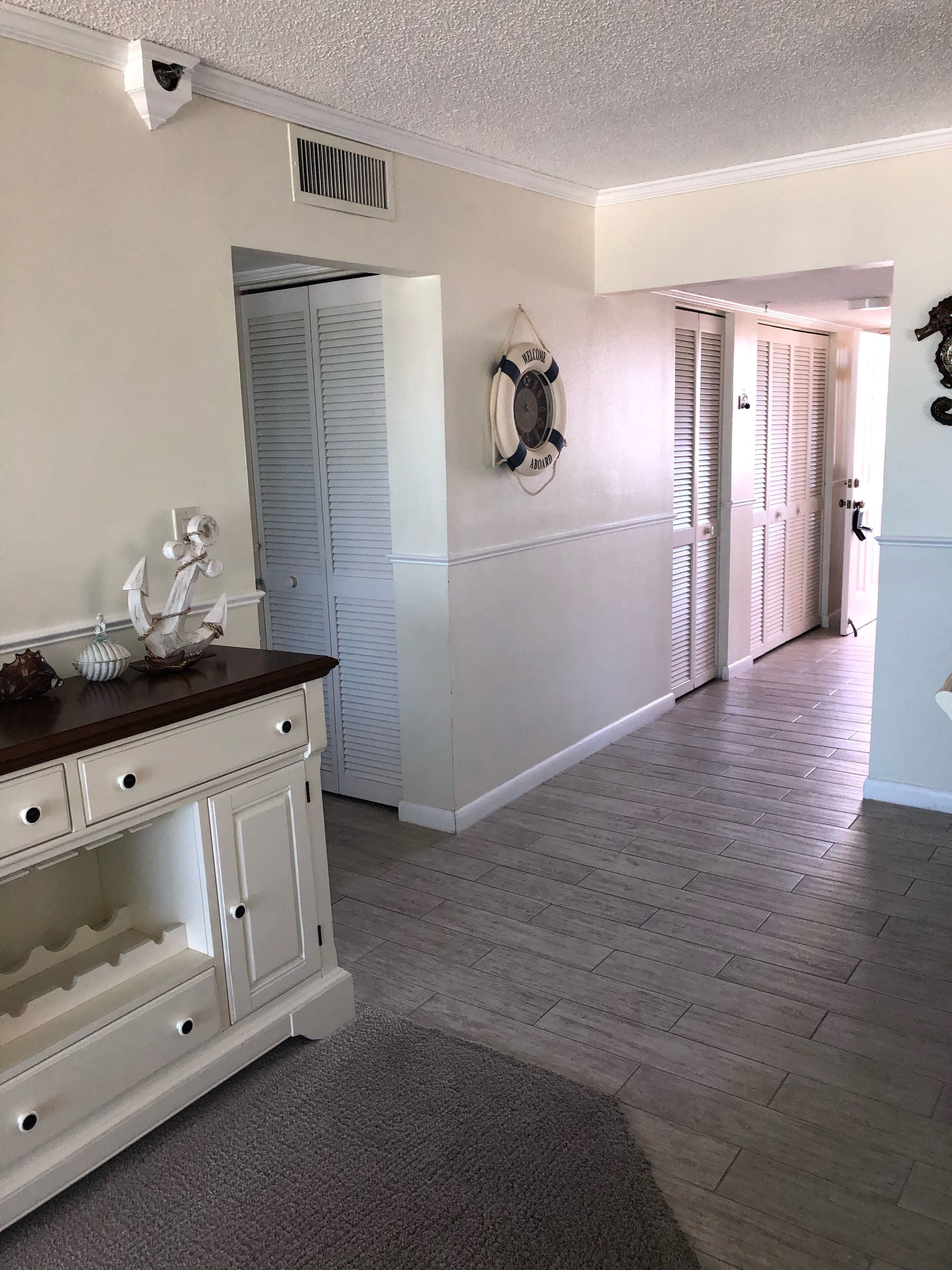 A hallway with a wooden floor, white walls, and a white door with shutters. There is a life preserver hanging on the wall and a small decorative anchor on the wall.