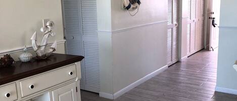 A hallway with a wooden floor, white walls, and a white door with shutters. There is a life preserver hanging on the wall and a small decorative anchor on the wall.
