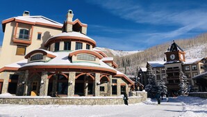Ein großes, mehrstöckiges Hotel mit schneebedecktem Dach und Steinfassade, mit einem markanten Glockenturm und einer Skipiste im Hintergrund.