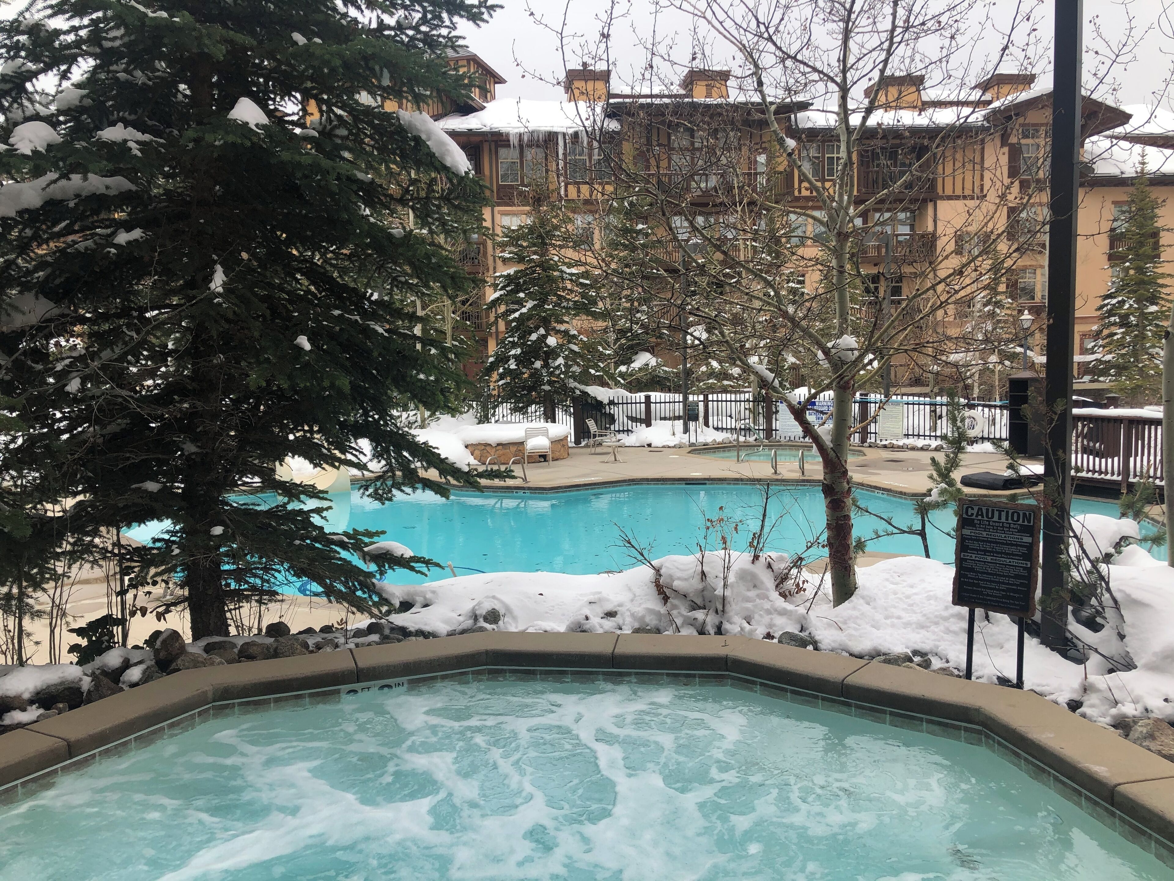 A pool area with a hot tub, surrounded by snow-covered trees and a building with balconies.