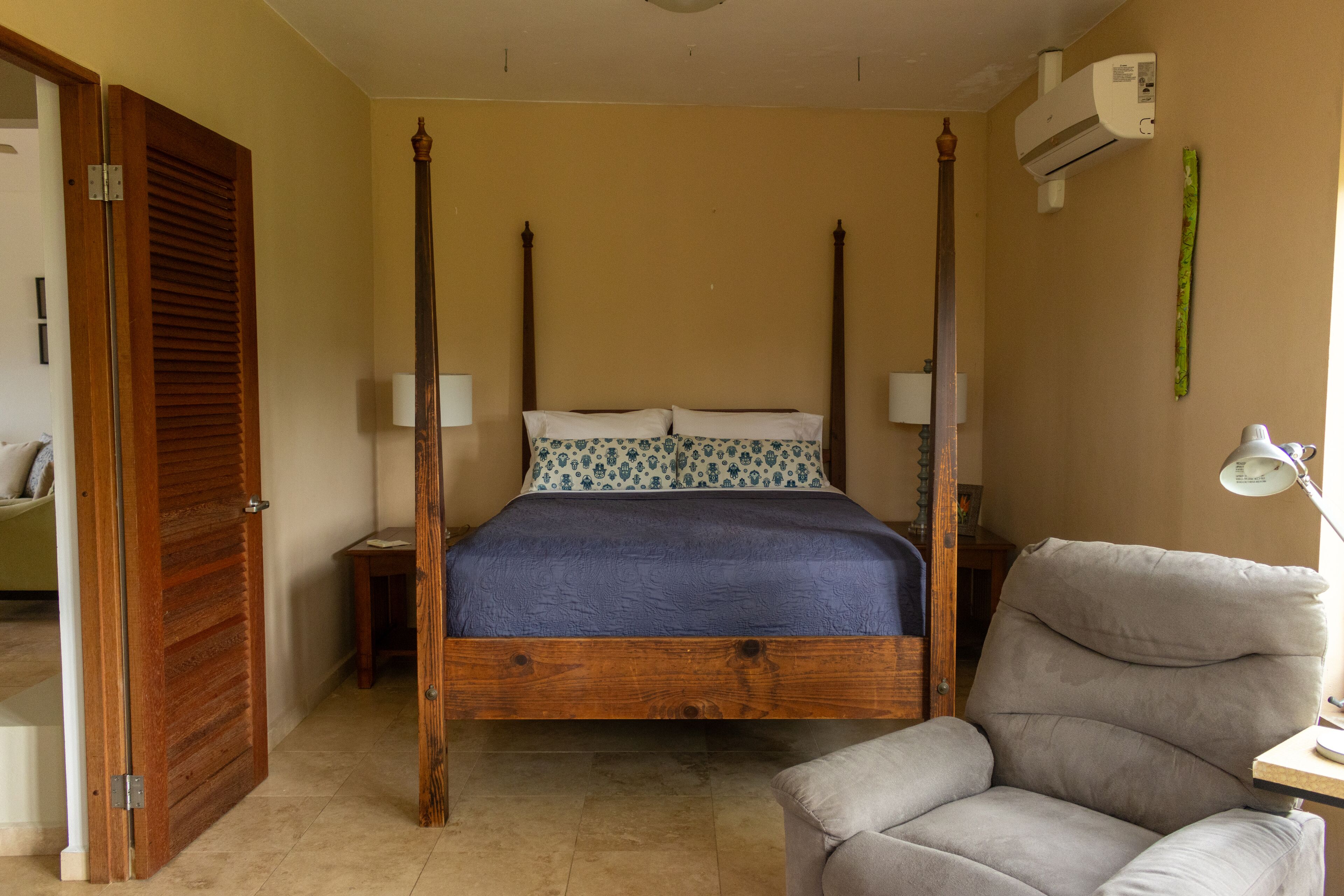 Un lit à baldaquin avec une couvre-lit bleue, deux tables de chevet avec des lampes, une chaise en bois et un climatiseur fixé au mur.