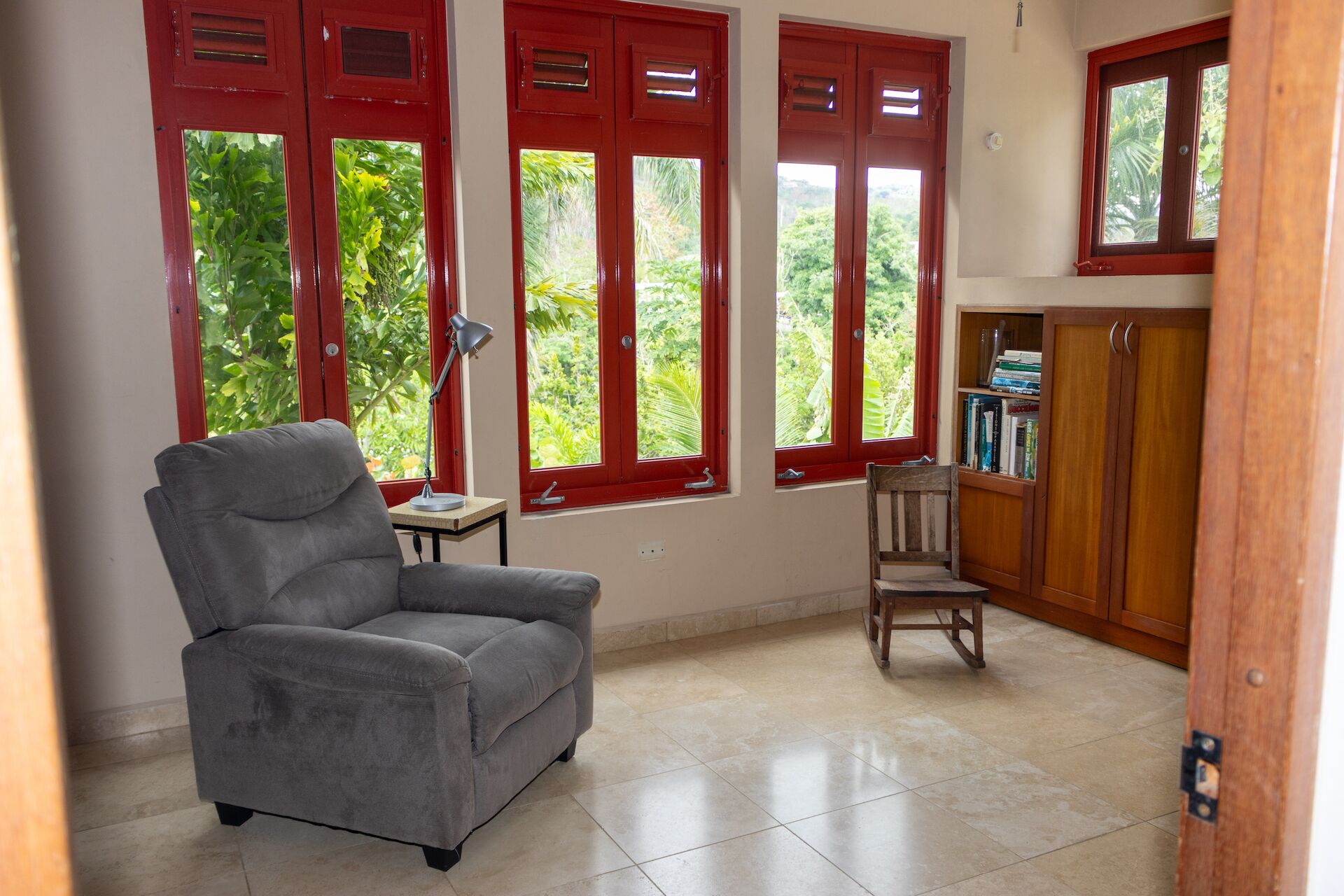 Una habitación con un sillón gris, una silla de madera, una mesita con una lámpara y una estantería.