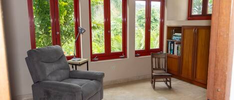 Una habitación con un sillón gris, una silla de madera, una mesita con una lámpara y una estantería.