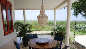 Eine überdachte Terrasse mit einem runden Holztisch und Stühlen, einer hängenden Leuchte und Blick auf Grünflächen und das Meer.