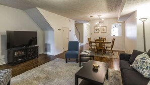 A living room with a flat-screen TV, dark wood flooring, a white shag rug, a dark sofa, a coffee table, and a dining area with a table and chairs. - A Renovated 3 Bedroom, 2.5 Bath Townhouse In Downtown Lexington. (Lexington)