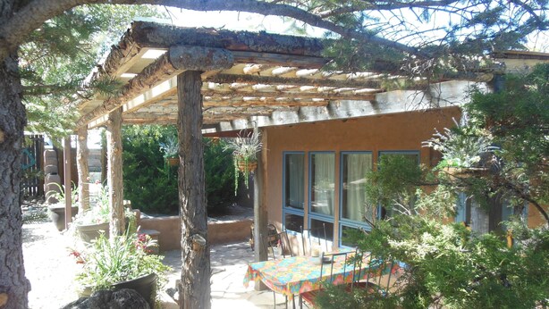 Un espacio al aire libre techado con mesa y sillas, rodeado de plantas en macetas y un banco de piedra.