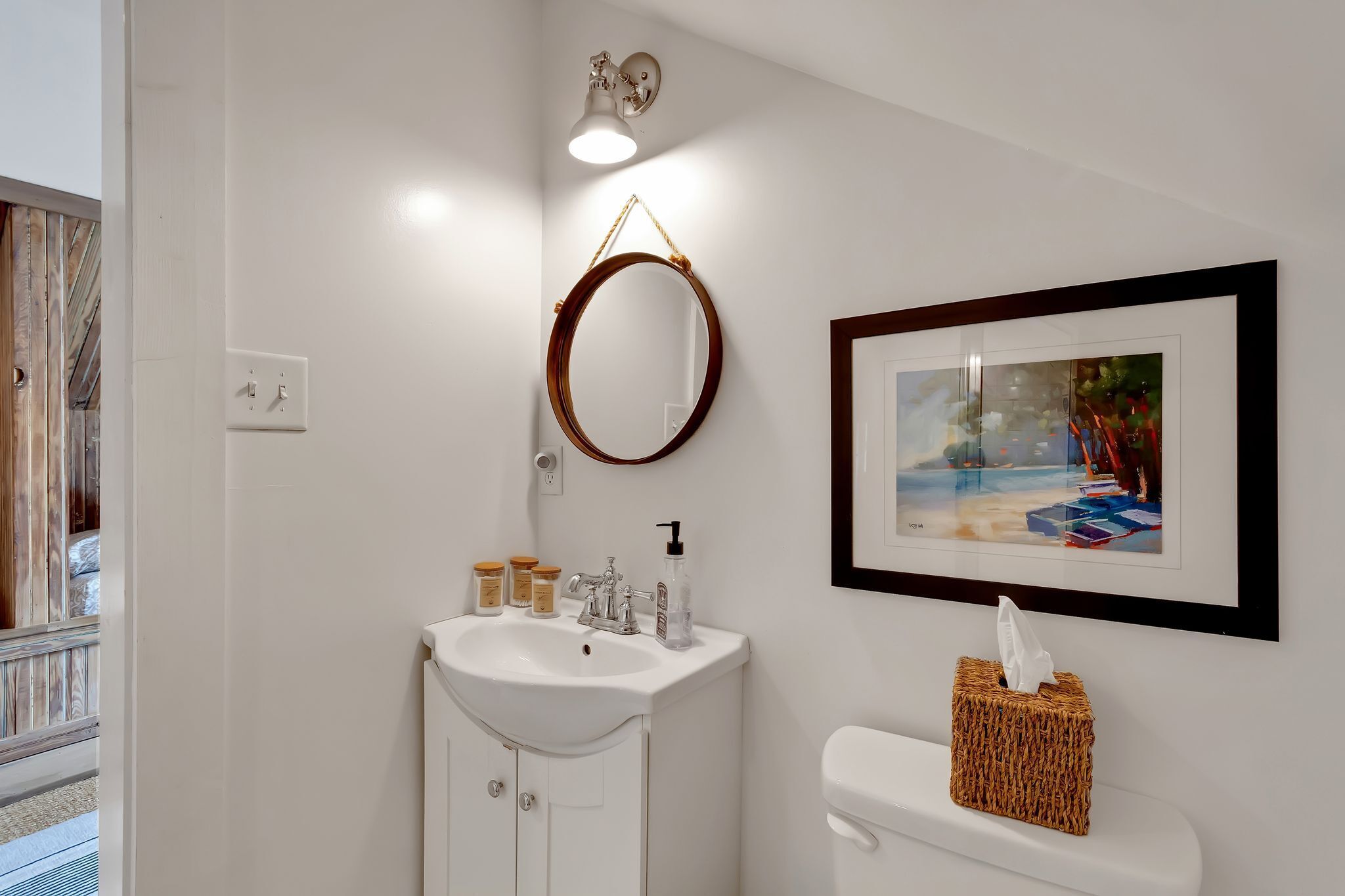 Un baño con un lavamanos blanco, un inodoro, un cuadro en la pared y una pequeña cesta de mimbre.