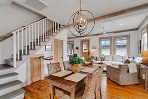 Un vaste espace de vie agrémenté d’un escalier en bois, d’une table à manger avec des sets de table en fibres tressées, de meubles en osier et d’un grand lustre.
