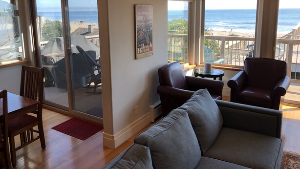 A spacious living room with a large window overlooking a beach, a comfortable gray sofa, and a wooden dining table. - Allegra Villa Alla Spiaggia  - See & Hear Ocean - Luxurious (Santa Cruz)