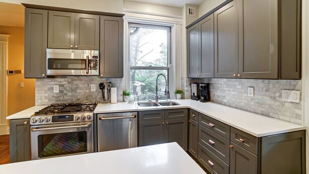A modern kitchen with grey cabinets, stainless steel appliances, a white countertop, and a window providing natural light. - Queen Ann Historic Mansion (Covington)