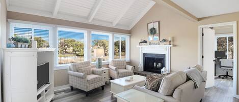 A spacious living room with a high ceiling, large windows, a fireplace, and a television.