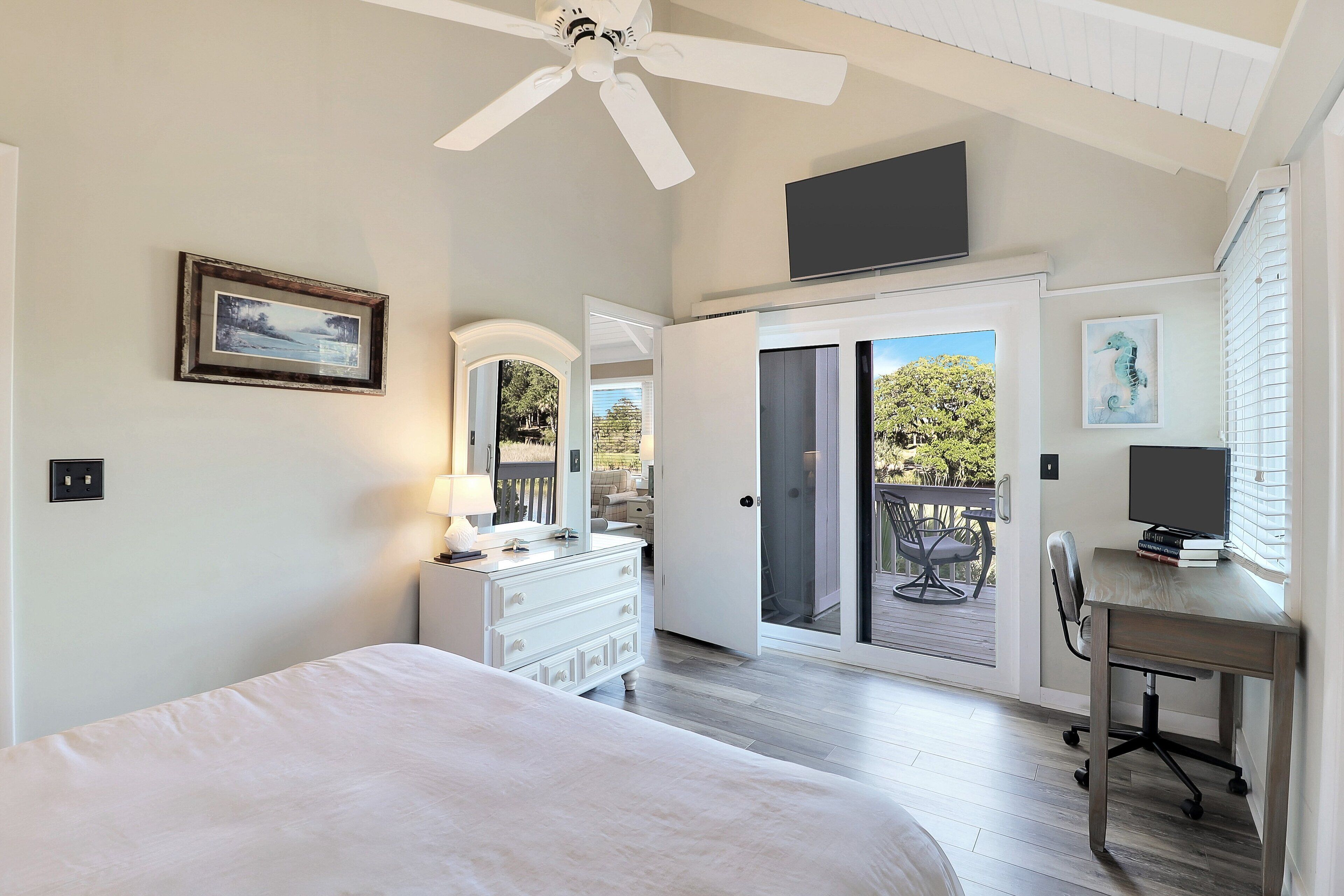 A bedroom with a bed, dresser, desk, and a ceiling fan. There is a sliding glass door leading to an outdoor area with a table and chairs.
