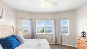 Un dormitorio con cama, ventilador de techo, dos ventanas con cortinas, cómoda y vista al exterior.