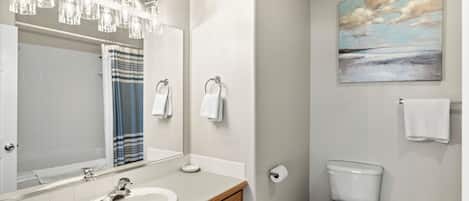 Un baño con lavamanos blanco, tocador de madera, un espejo grande, cabina de ducha de vidrio, inodoro y un cuadro en la pared.