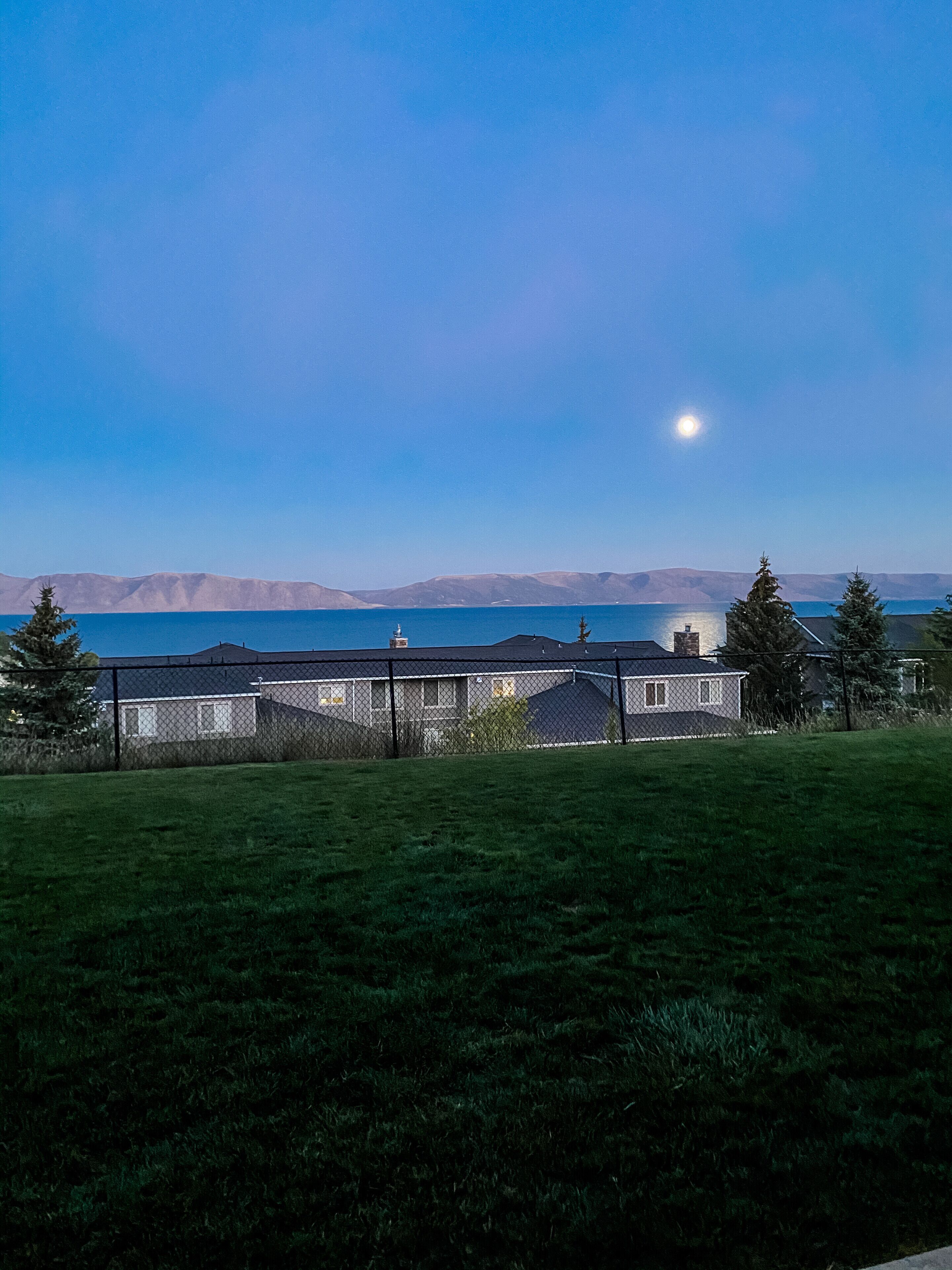 Un barrio residencial con casas, un campo cubierto de césped y una vista despejada de un lago y montañas a lo lejos.