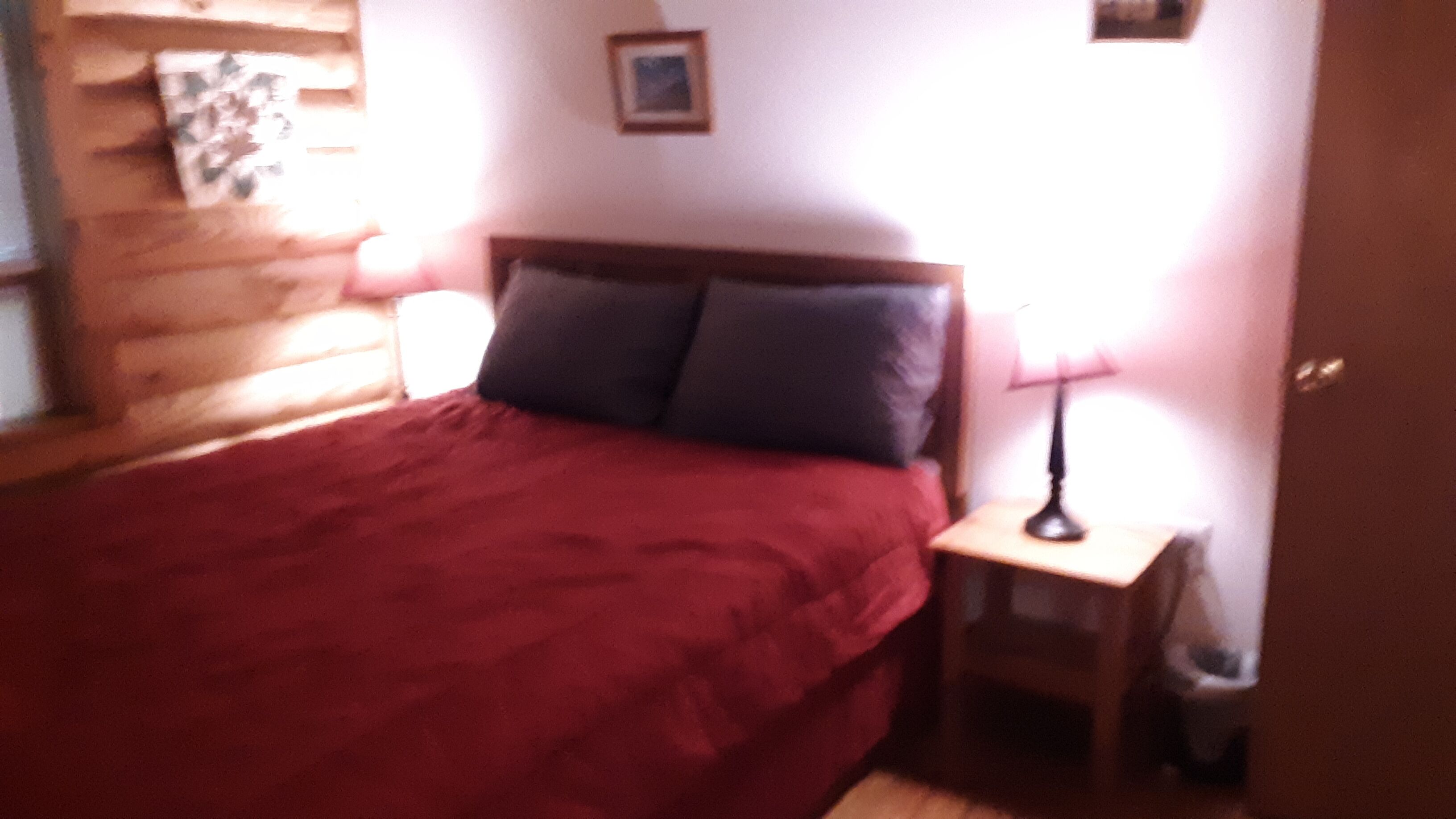 Ein Schlafzimmer mit einem Holzkopfteil, einer roten Bettdecke, zwei Kissen, einem Nachttisch mit einer Lampe und einem kleinen Bild an der Wand.