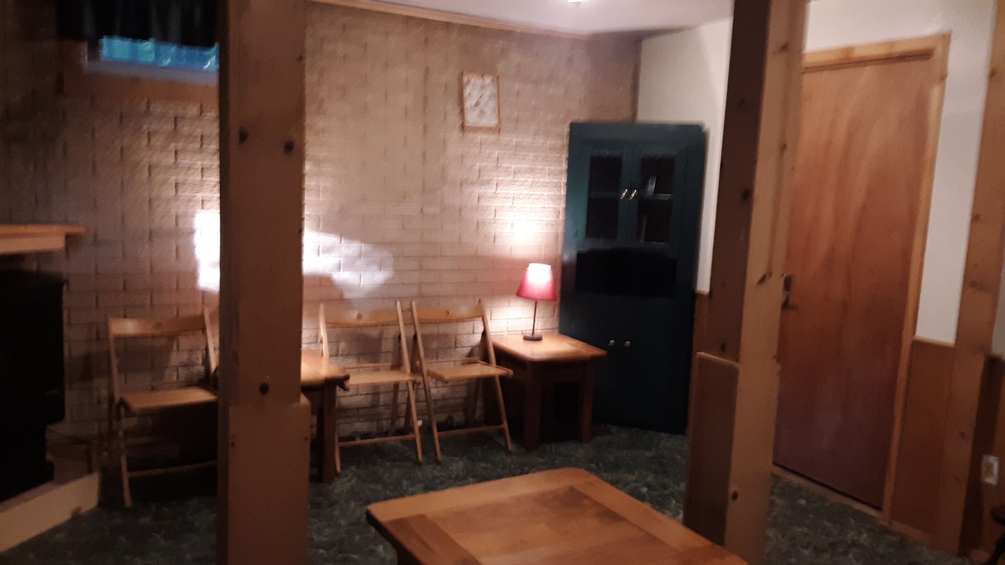 A room with a brick wall, a wooden table, a chair, a lamp, and a door.