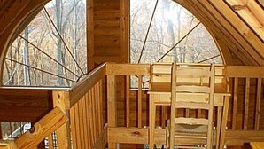 Une cabane en bois avec une chambre à coucher située en mezzanine, un escalier en bois et une vue sur les arbres, visible à travers de grandes fenêtres en arc de cercle.