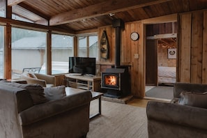 A cozy living room with a fireplace, comfortable seating, a television, and large windows. - Rustic Lakefront Cabin with Bonus 'Tree House' - A Place to Unwind (Port Angeles)
