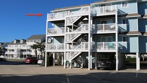 A multi-story residential building with balconies, a parking area, and a clear blue sky. - Monkey Sea, Monkey Dune ~ Top floor, End unit; Sound and Ocean view (Ocean Isle Beach)