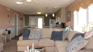A living room with a beige sofa, patterned pillows, a ceiling fan, and a view into the kitchen. - Monkey Sea, Monkey Dune ~ Top floor, End unit; Sound and Ocean view (Ocean Isle Beach)