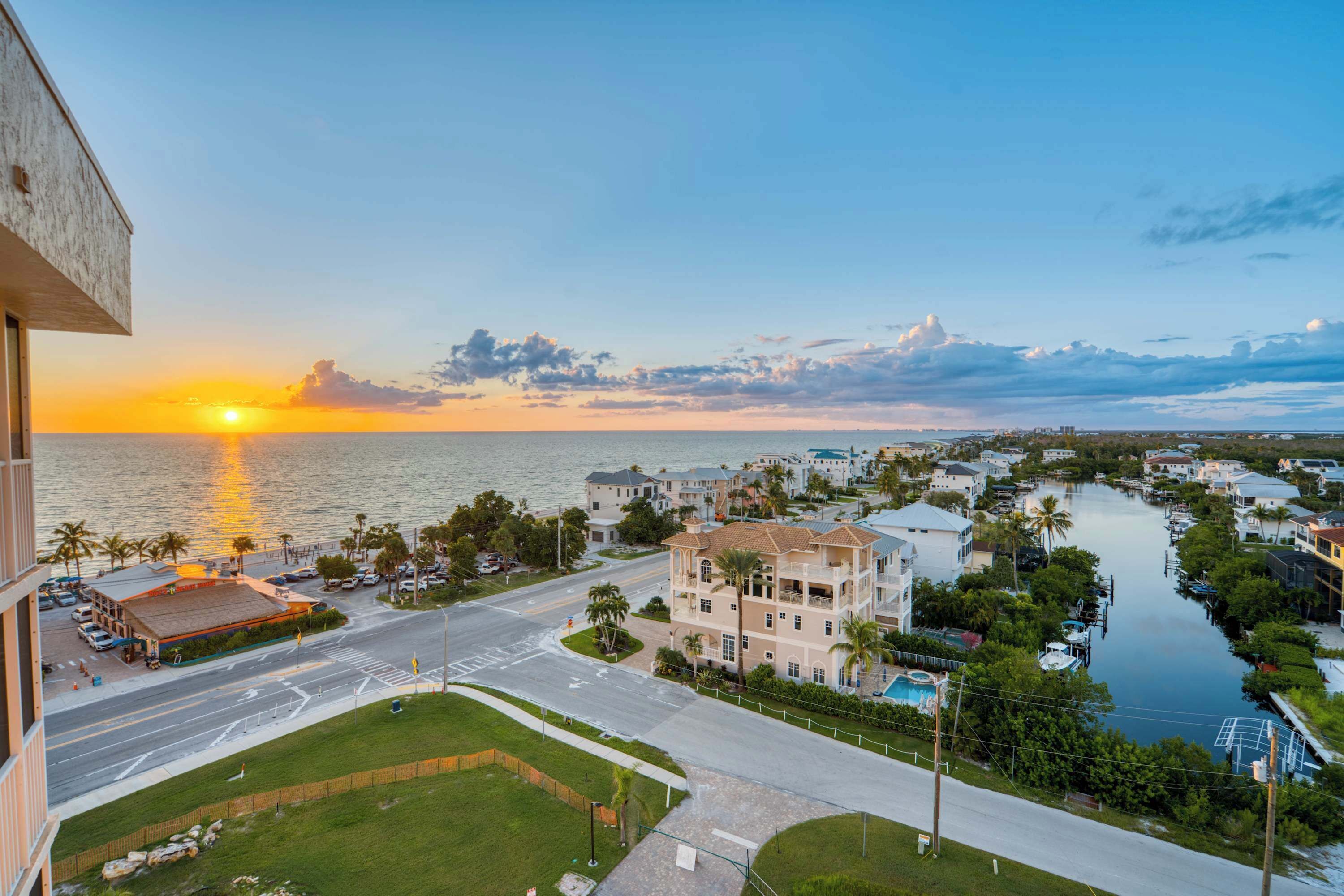 Un paisaje urbano costero con un atardecer sobre el agua, que muestra una combinación de edificios residenciales y comerciales, un puerto deportivo y una carretera despejada que conduce al paseo marítimo.