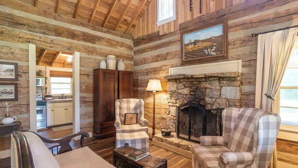 A log cabin interior with a fireplace, checkered armchairs, a sofa, and a kitchen area visible through an open doorway. - The Pines Cabin at Green Frog Farm (Bells)