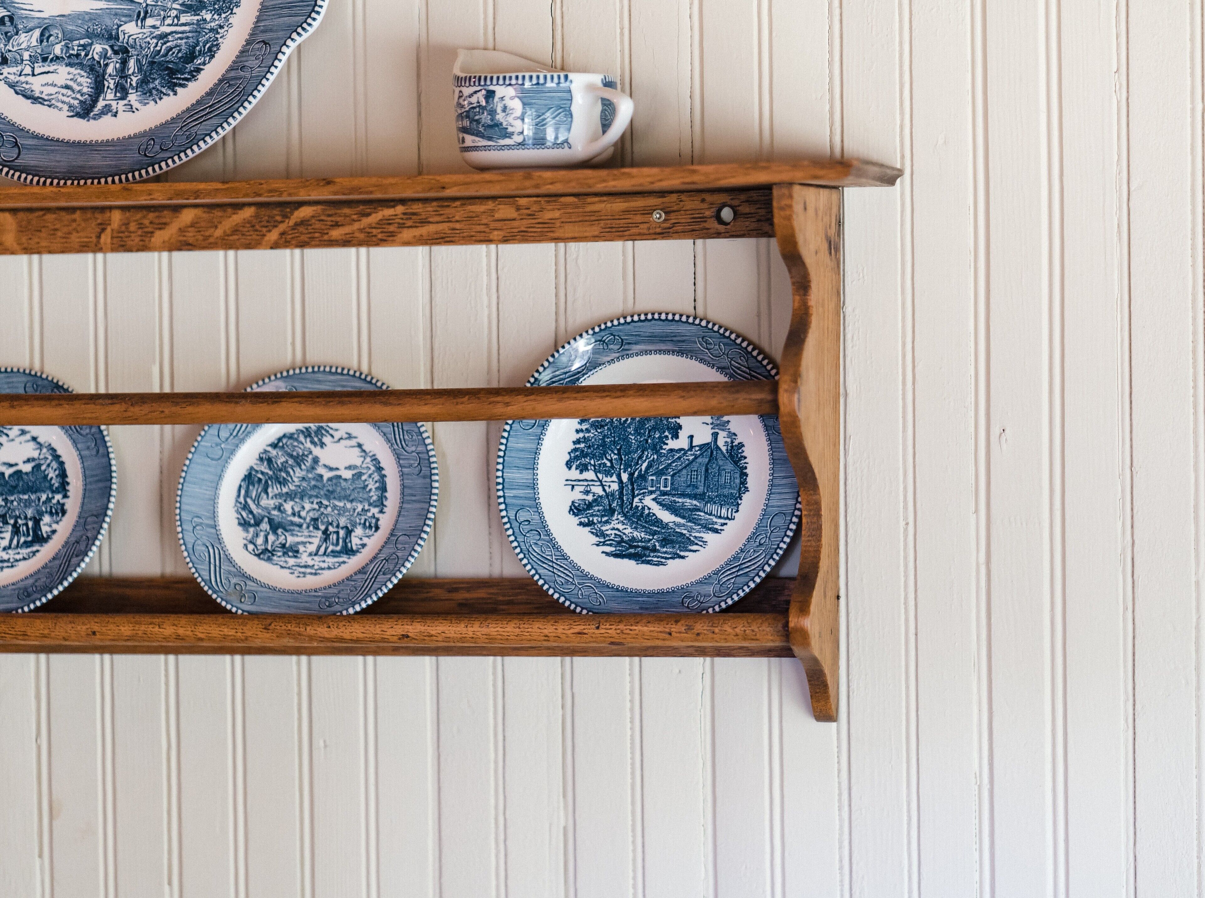 A wooden shelf with four blue and white plates and a matching cup, against a white paneled wall.