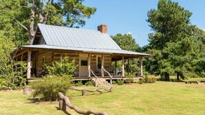 A rustic log cabin with a metal roof, surrounded by a grassy field and trees. - The Pines Cabin at Green Frog Farm (Bells)