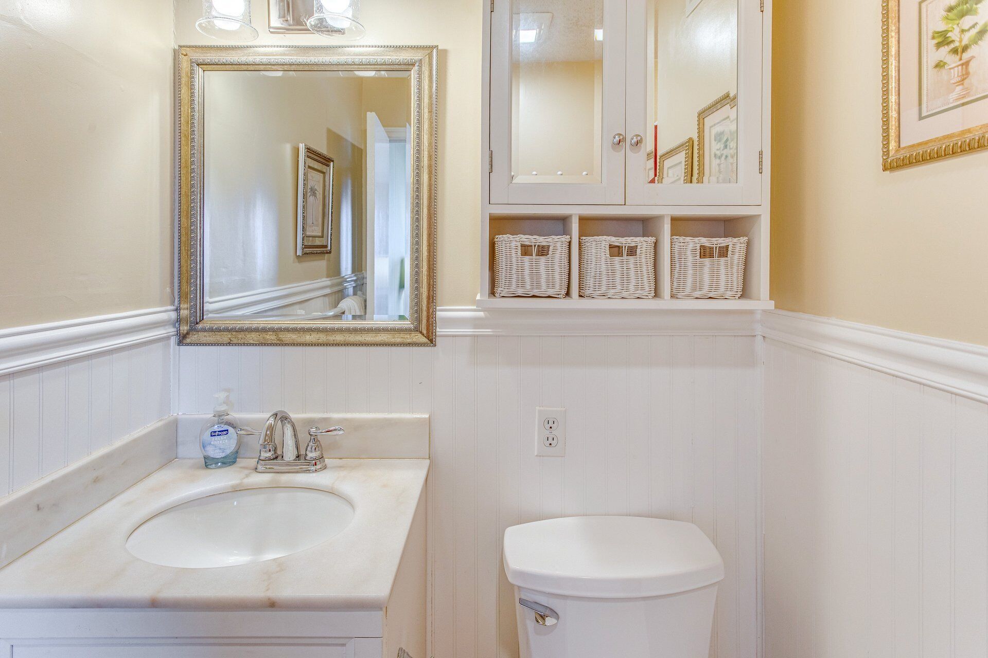 A bathroom with a double sink, a toilet, a mirror with an ornate frame, and wicker baskets on the wall.
