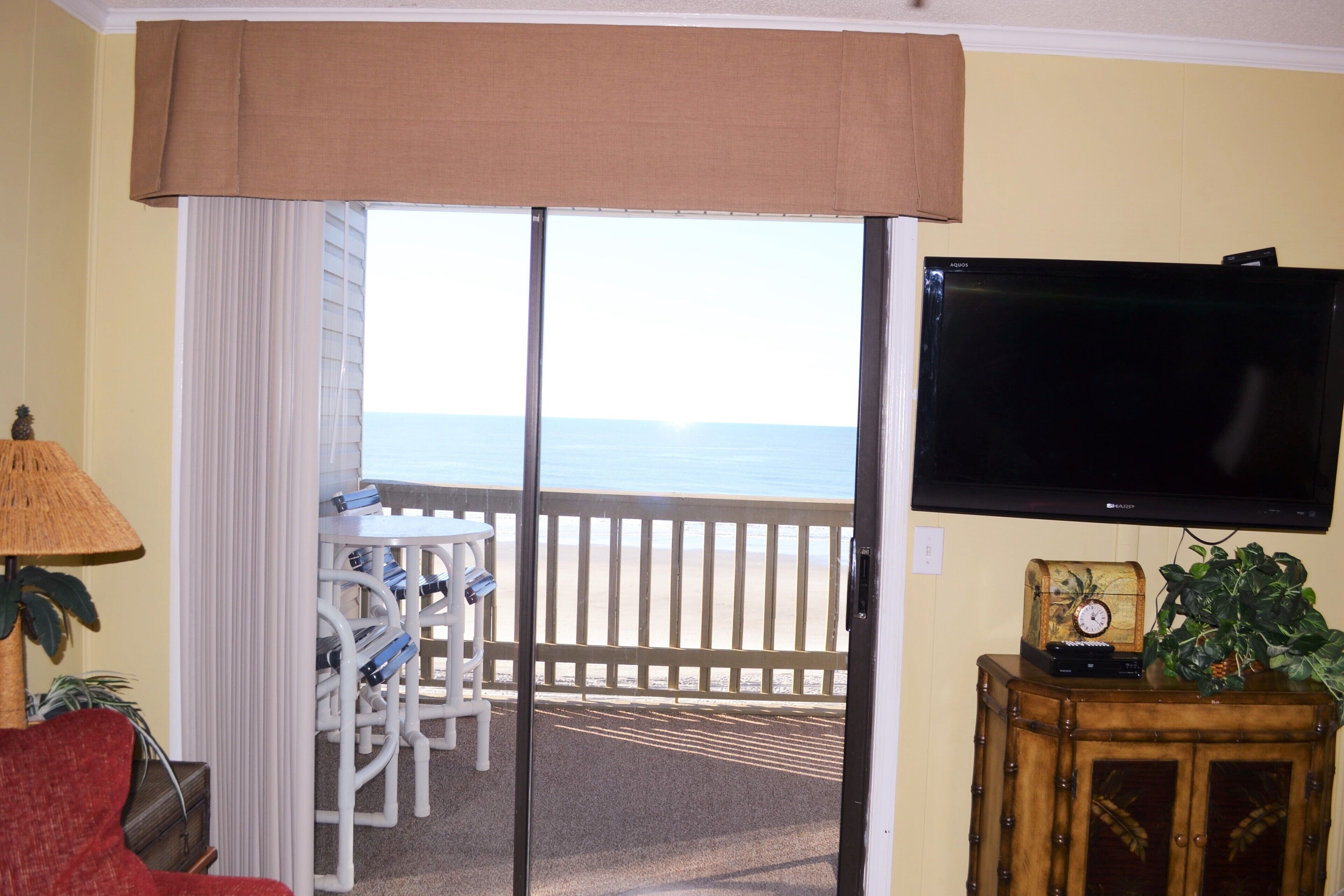 Habitación con puerta de cristal corrediza que da a un balcón con vista al océano. Se ve un televisor montado en la pared, un pequeño mueble de madera con un reloj y una planta sobre él.