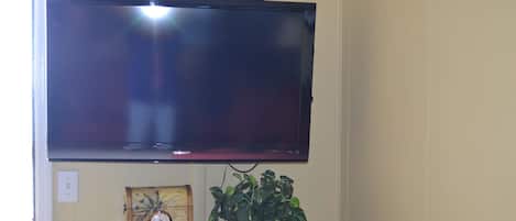 A television mounted on a wall, a wooden side table with a clock and a plant, and a power outlet.