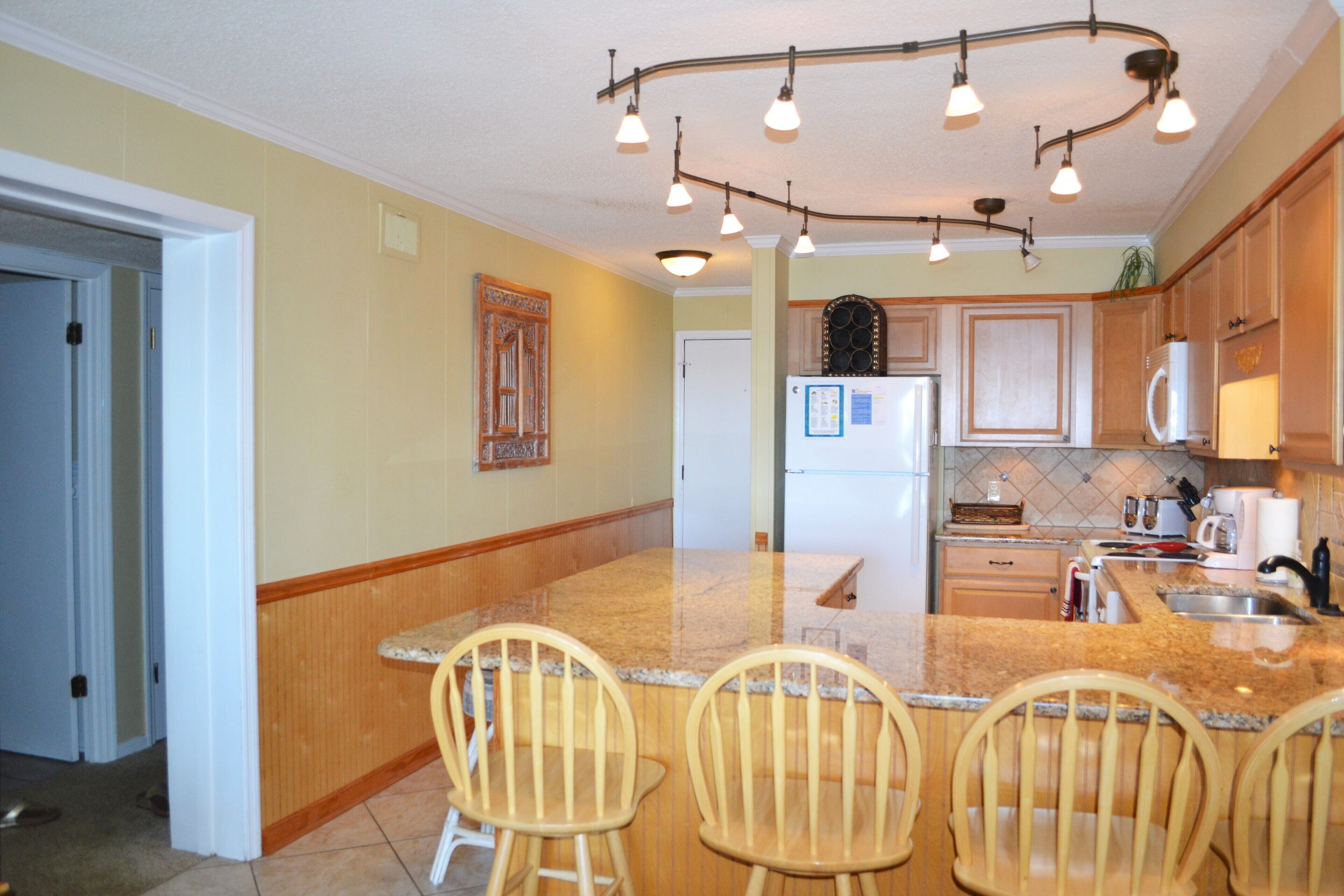 A kitchen with a granite countertop, wooden chairs, a refrigerator, and a microwave.