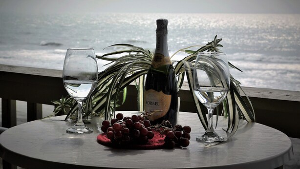 A table with a bottle of wine, a glass of water, and a bunch of grapes on a balcony overlooking the sea.