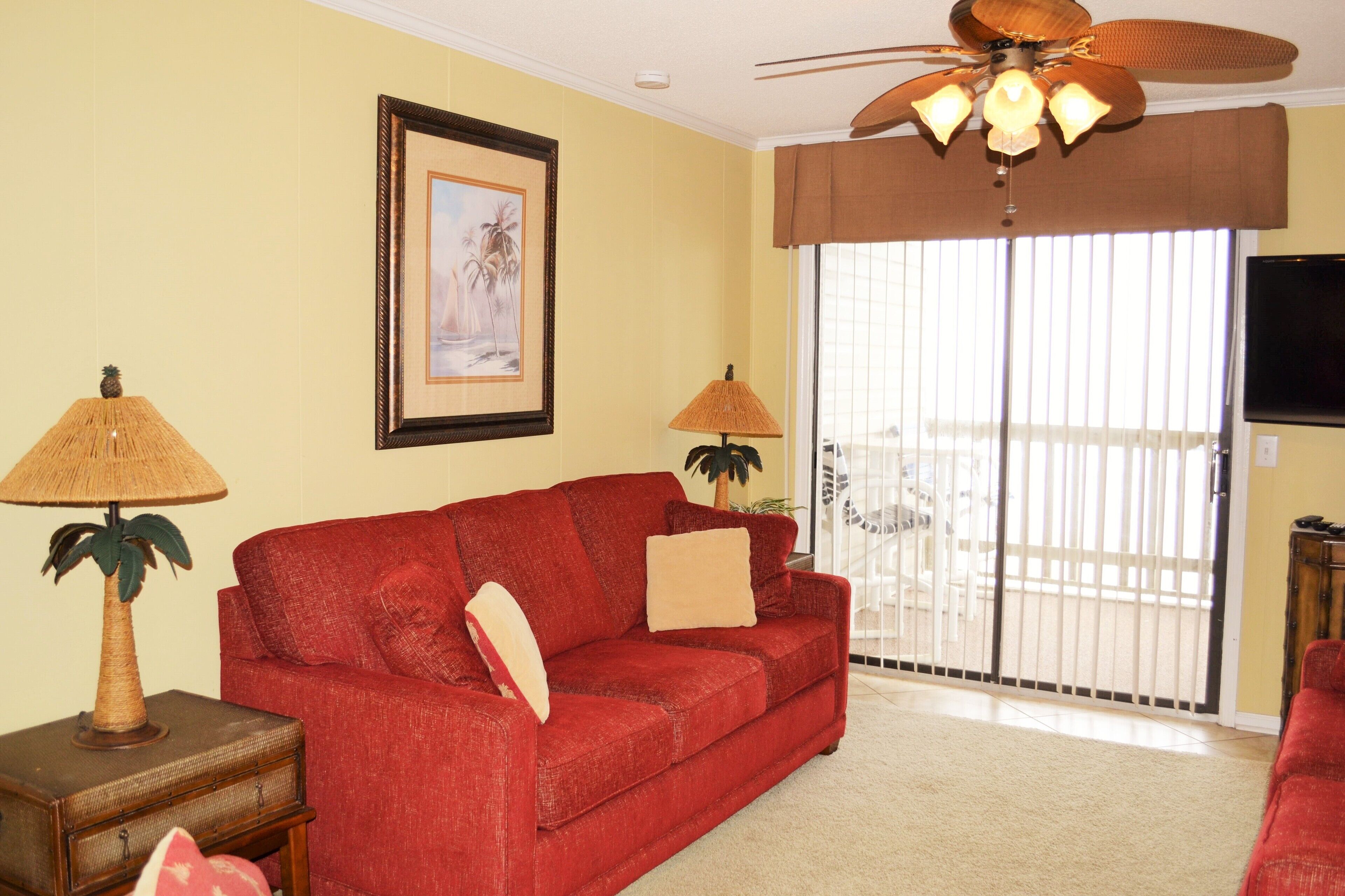 A living room with a red sofa, two lamps, a ceiling fan, a framed picture, and a sliding glass door.