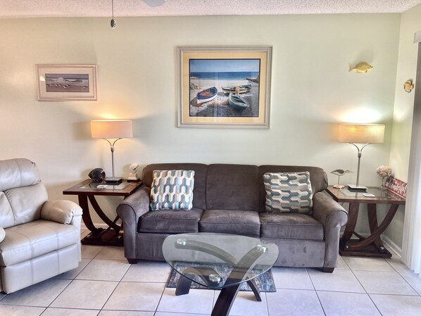 A living room with a grey sofa, a glass coffee table, and two lamps. There is a framed picture of boats on the wall. - OCEAN VIEW Villa. Steps to the Beach! Pool, Hot Tub, SPECIAL OFF-SEASON RATES! (St Augustine)