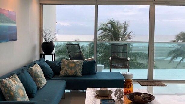 Una sala de estar moderna con un amplio ventanal con vistas al mar, un sofá azul con cojines estampados, una mesa de centro con objetos decorativos, y una vista de palmeras y el océano.